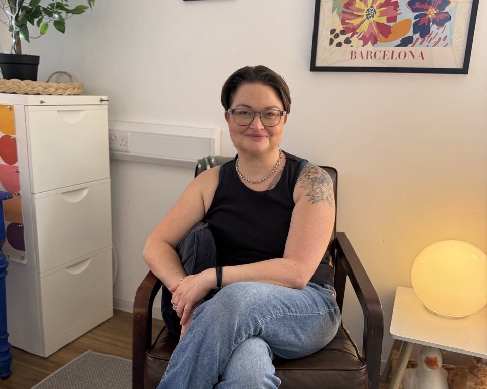 Delia sitting in the armchair in her counselling room, with a lamp lit beside her.