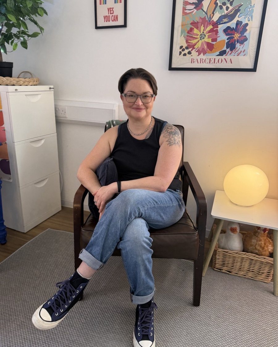 Delia, sitting in her counselling room in a black top and jeans, smiling at the camera.