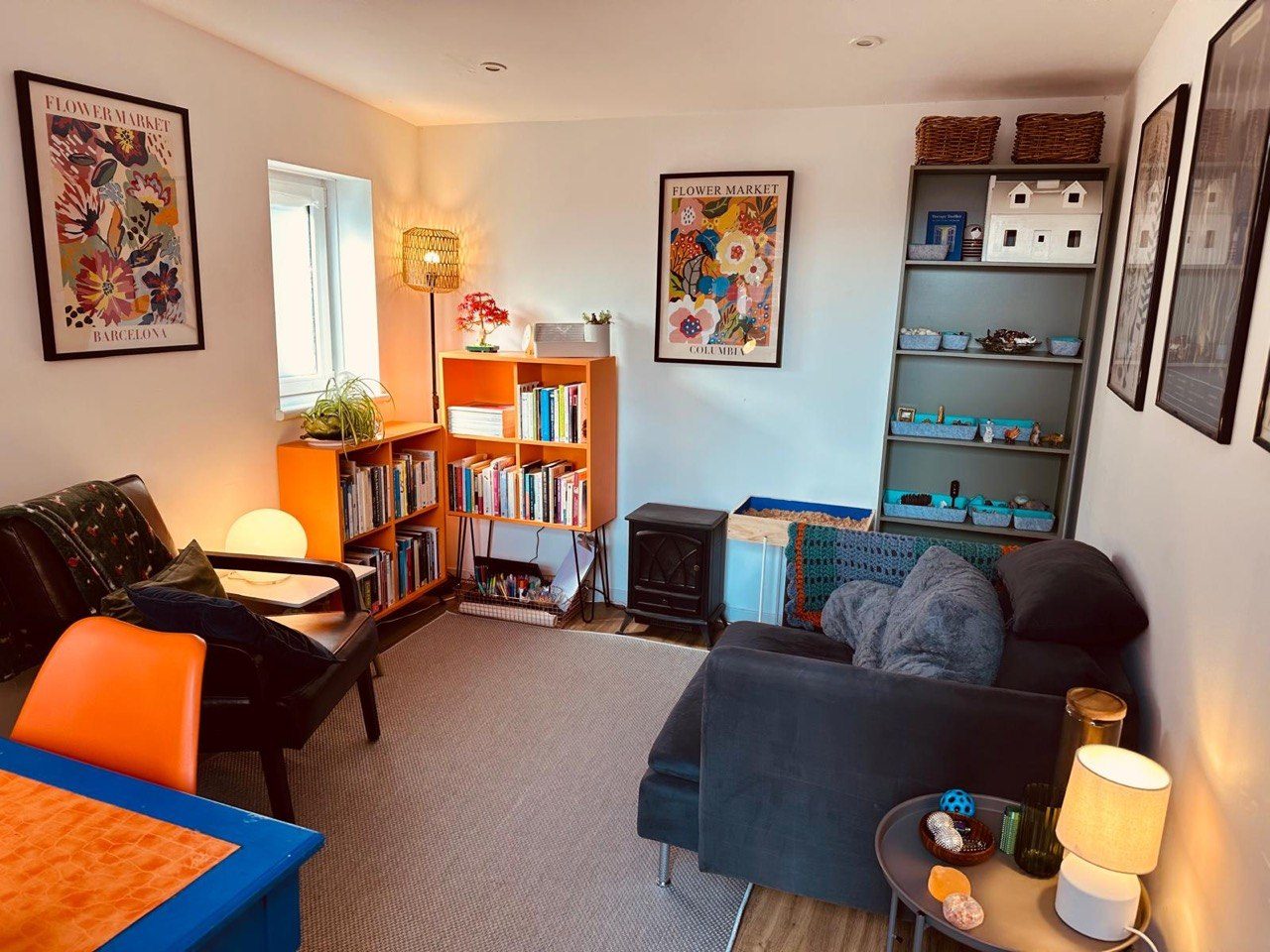 Inside the counselling room: a grey sofa with cushions and blankets, orange shelving, plants and framed prints.